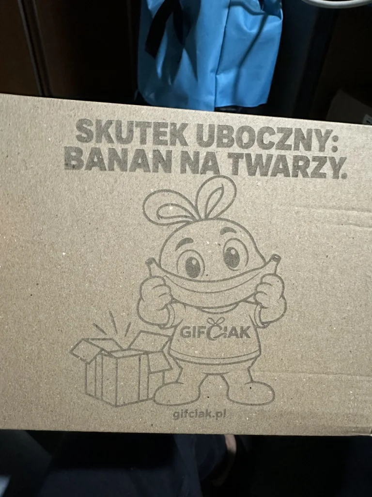 Karton Gifciak z bliska: wygrawerowany Gifciak z bananem zamiast uśmiechu, otwarte pudełko i napis „Skutek uboczny: banan na twarzy”